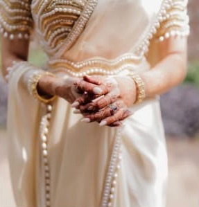 Hands clasped over an ivory saree with a pearl-trimmed border