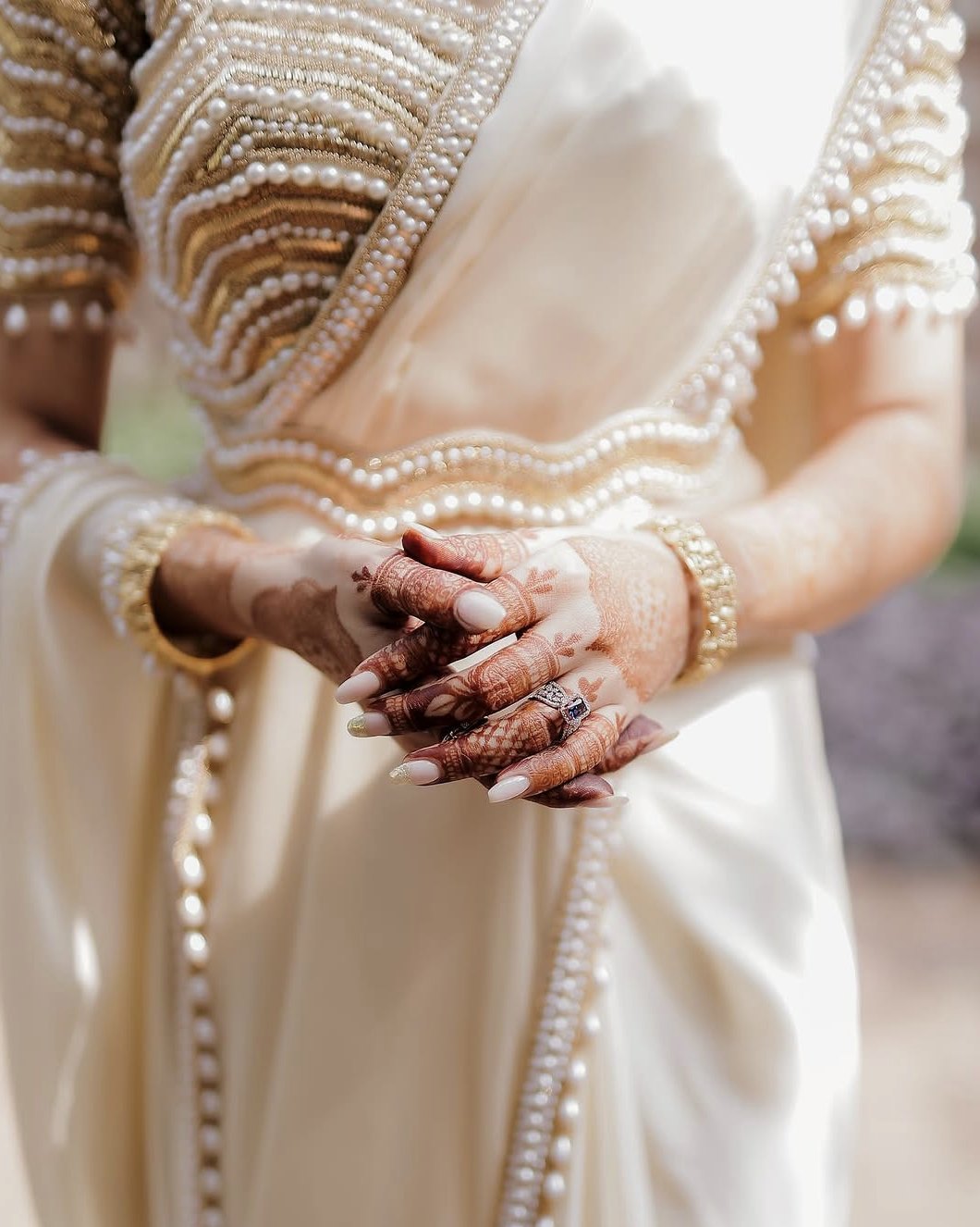 Hands clasped over the ivory saree, a pearl-trimmed border and gold bangles